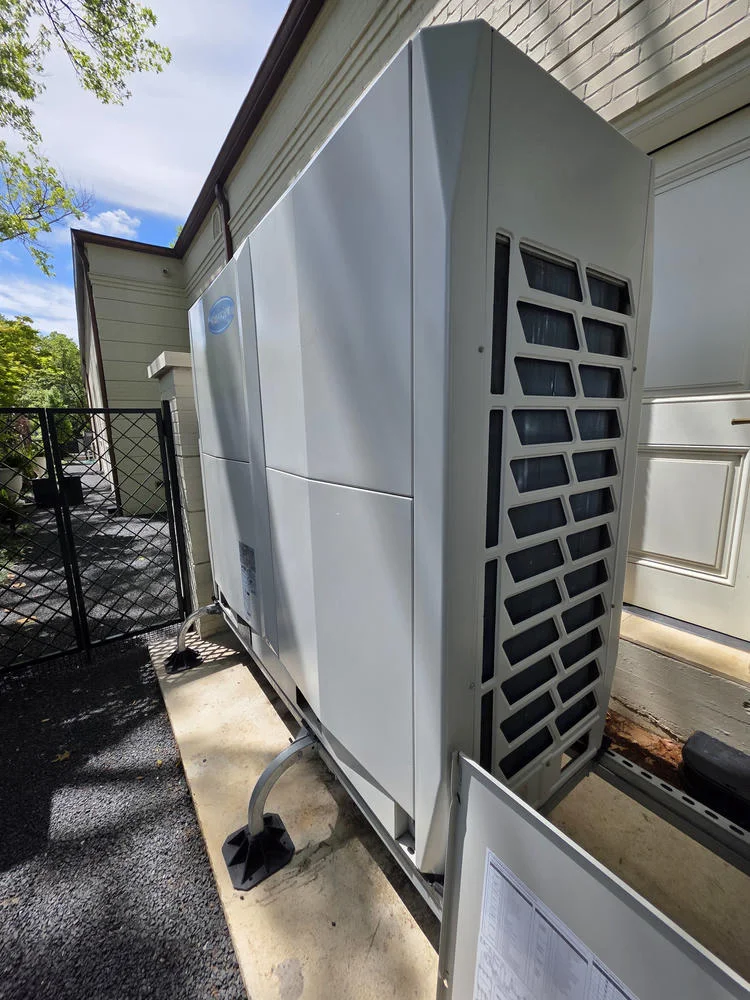 A large, gray Carrier air conditioning unit sits next to a beige brick building, with a black fence and gravel.